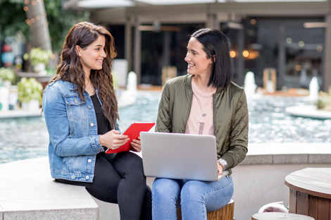 Two women sitting outside with a tablet and laptop, talking