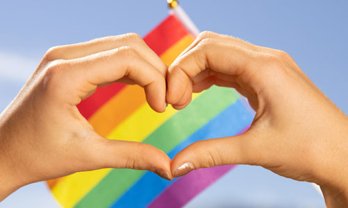 Hands shaped like a heart with rainbow flag in background