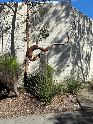 a sculpture made of steel that has partly rusted supported by a timber block next to scrubs and trees