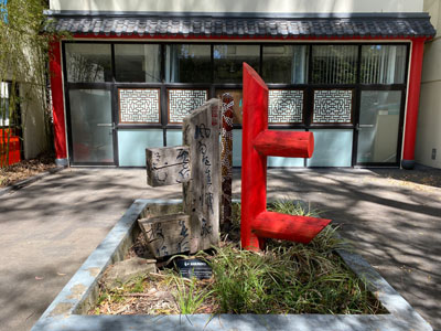 a sculpture made of two wooden structures with one on the right painted red in front of a chinese-inspired architecture