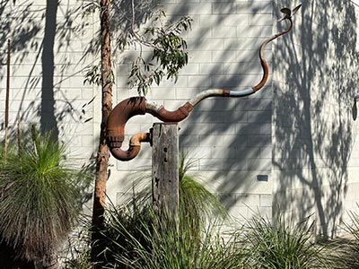 a sculpture made of steel that has partly rusted supported by a timber block next to scrubs and trees