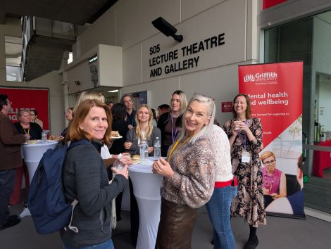 People chatting around a bar table at GCMH Research Showcase outside the Lecture Theatre, with Mental Health Wellbeing banners visible in the background.