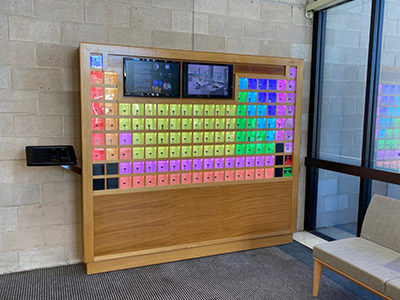 A 3-dimensional periodic table and two LCD screens on a large wooden structure leaning against the wall