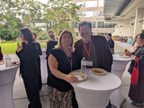 Two attendees stand beside a cocktail table GCMH event, with other guests, refreshments, and a building courtyard in the background.