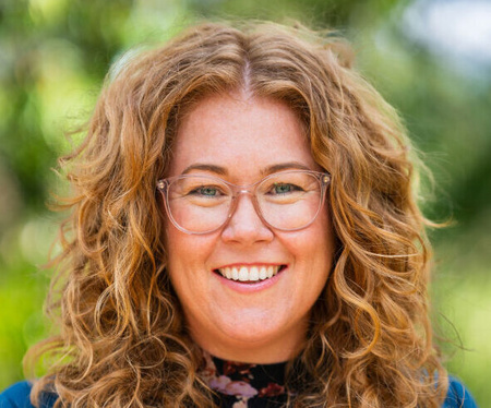 Woman with auburn curly hair, wearing glasses and smiling.