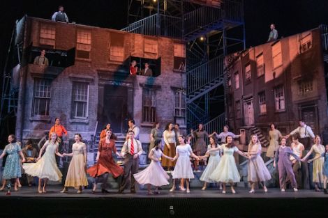 Group of musical theatre performers on a stage with a large building set behind them